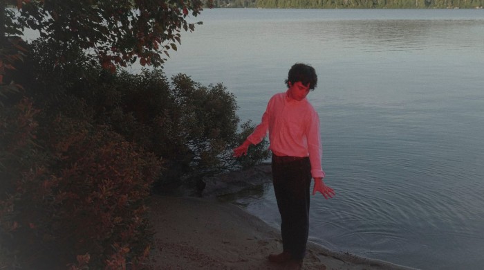A Man wearing a white long sleeve top, black trousers, brown belt, and brown shows, with curly dark brown hair stands at the edge of a lake on the sand with his back to the water. There is a red hued light shining towards him